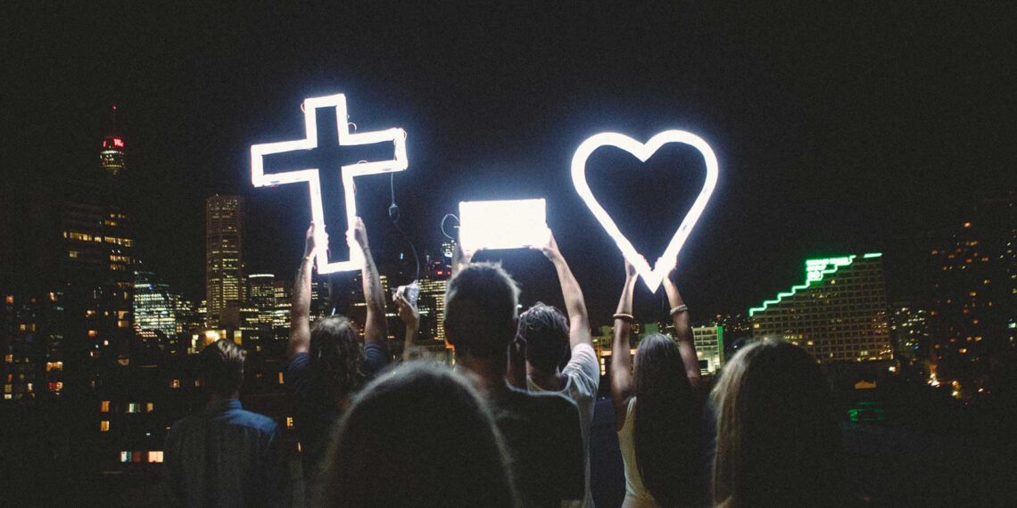 photo of crowd holding lighted 'cross equals love' signs against nighttime city metro background
