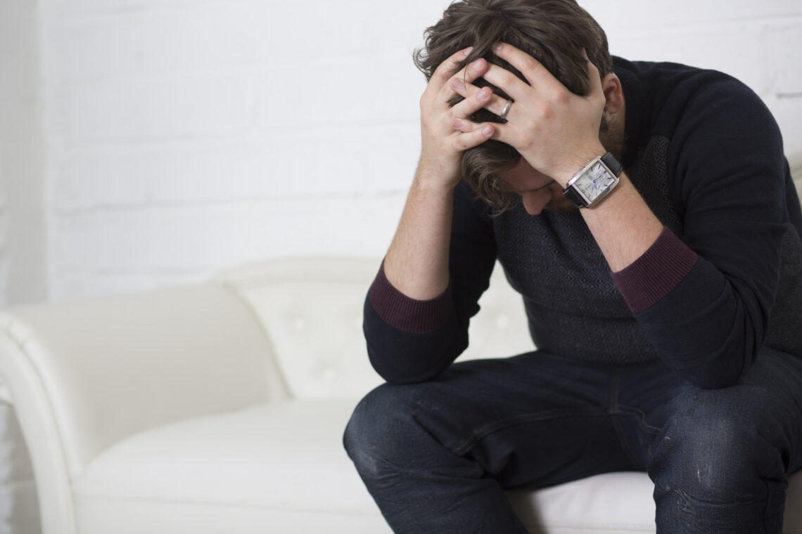 photo of man sitting on a white couch, upset and holding his head in his hands