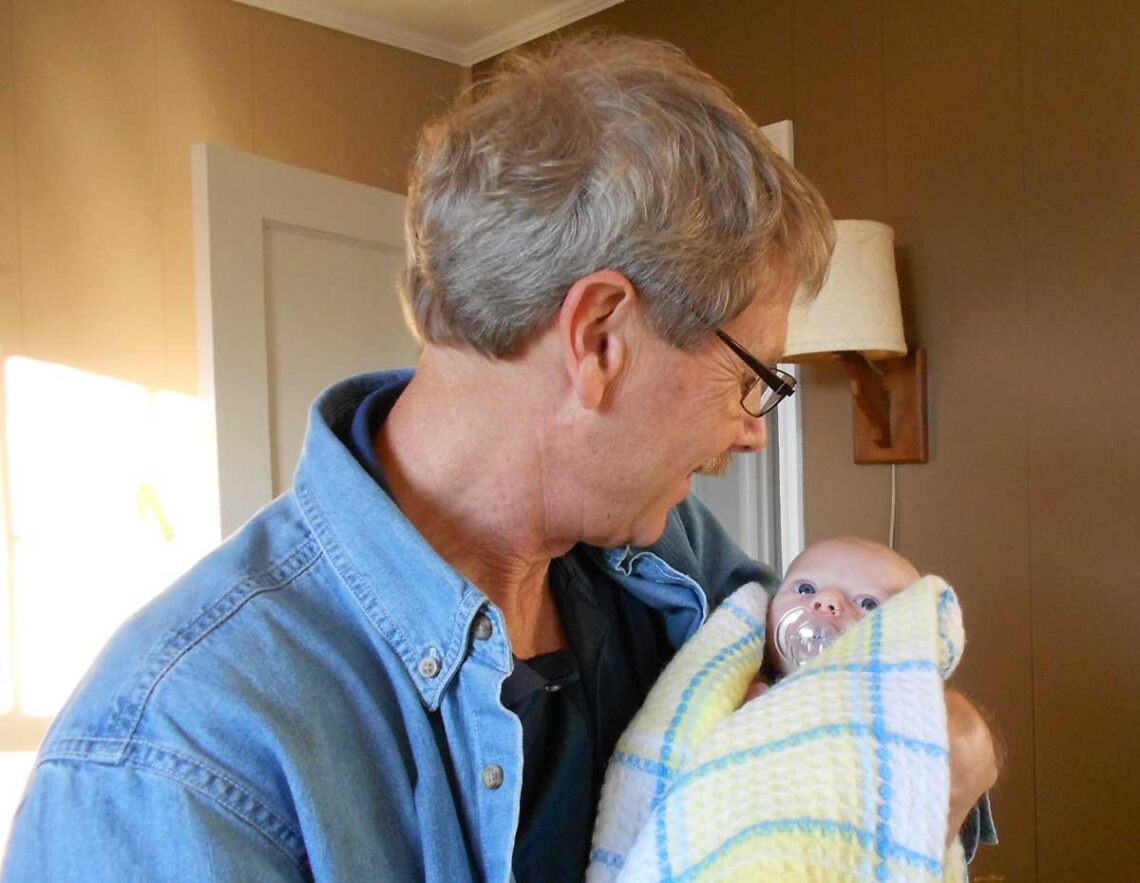 photo of older man with glasses in a denim shirt holding a newborn infant