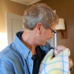 photo of older man with glasses in a denim shirt holding a newborn infant